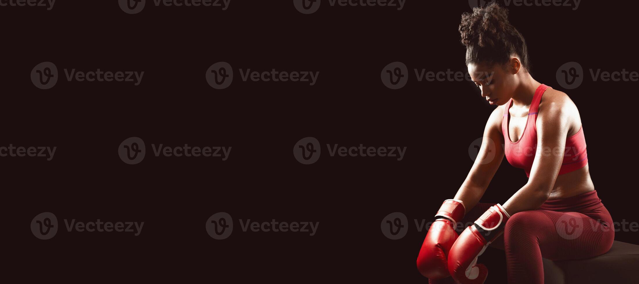 African muscular woman boxer getting ready for fight, sitting and looking down over black background, copy space photo