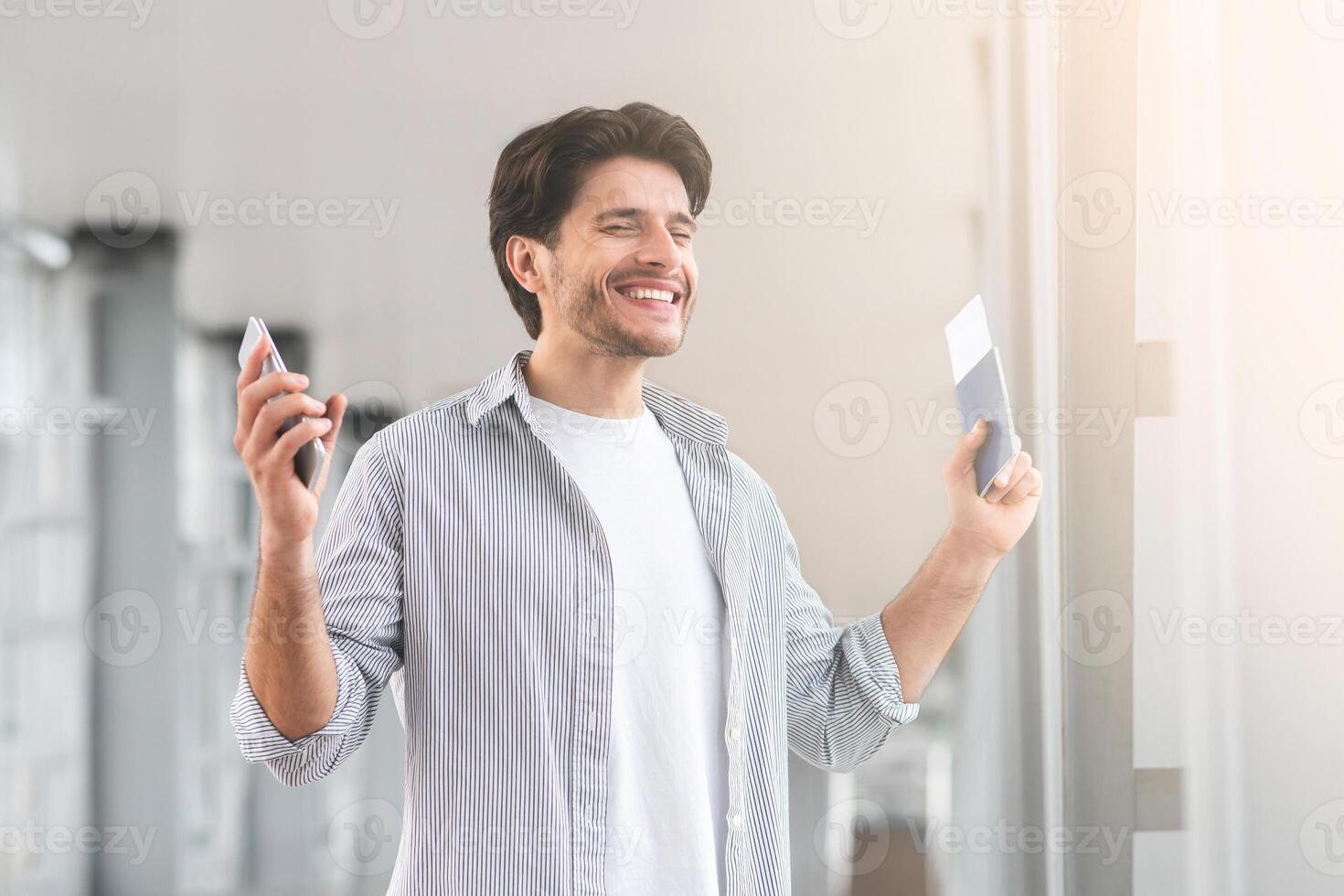 Enjoying Travels. Portrait Of Happy Young Man Standing In Airport, Holding Passport With Tickets And Smartphone In Hands, Raising Hands In Excitement photo