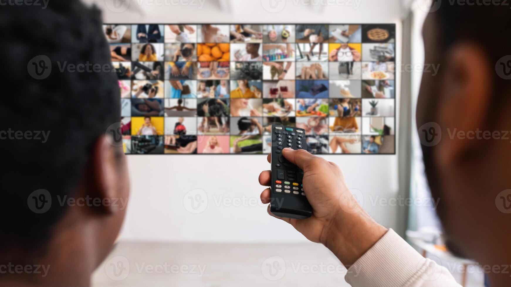 A man sits in front of a television screen that is displaying a grid of smaller screens, each showing a different image or . He is using a remote control to change the channel. photo