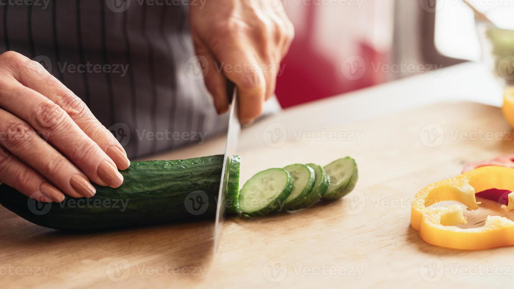 A woman hand slices a green cucumber on a wooden cutting board in a kitchen, with already sliced cucumbers and a sliced bell pepper in the background, cropped photo
