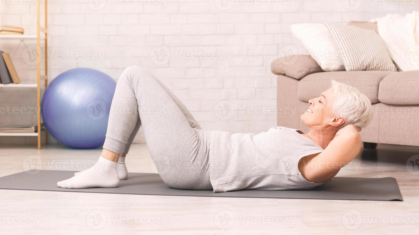 An older woman is engaging in a crunch exercise on a yoga mat, demonstrating strength and flexibility. Her focused expression shows dedication to her fitness routine. photo