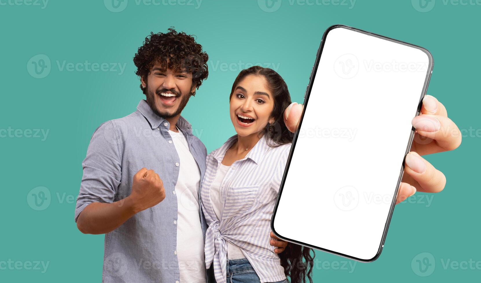 A young Indian couple stand against a teal background. They both look directly at the camera and smile. The woman holds a smartphone in front of them, displaying a blank white screen photo