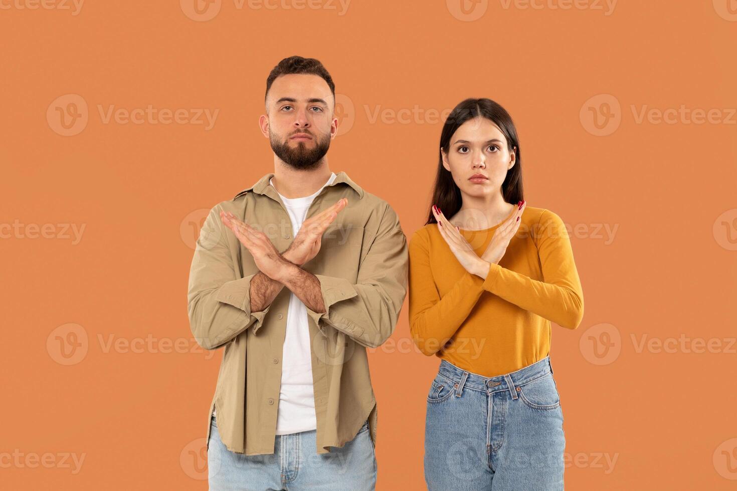 A young man and woman stand side by side against an orange background, displaying a clear no gesture by crossing their arms in front, appear serious, indicating disapproval or disagreement photo