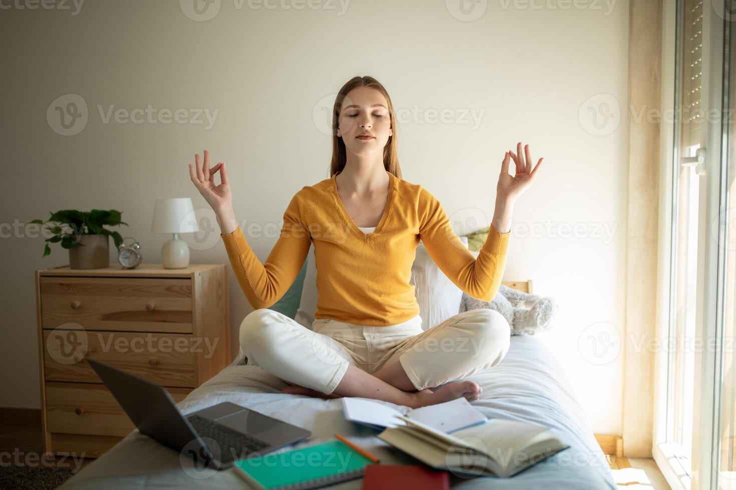 A teen girl is sitting on her bed, meditating with closed eyes and a serene expression. She is surrounded by open books, a laptop, and notebooks, indicating she is taking a break from studying photo