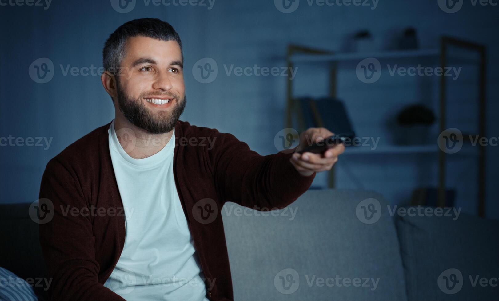 A bearded man sits on a couch, smiling while pointing a remote control towards a television in a cozy living room at night. photo
