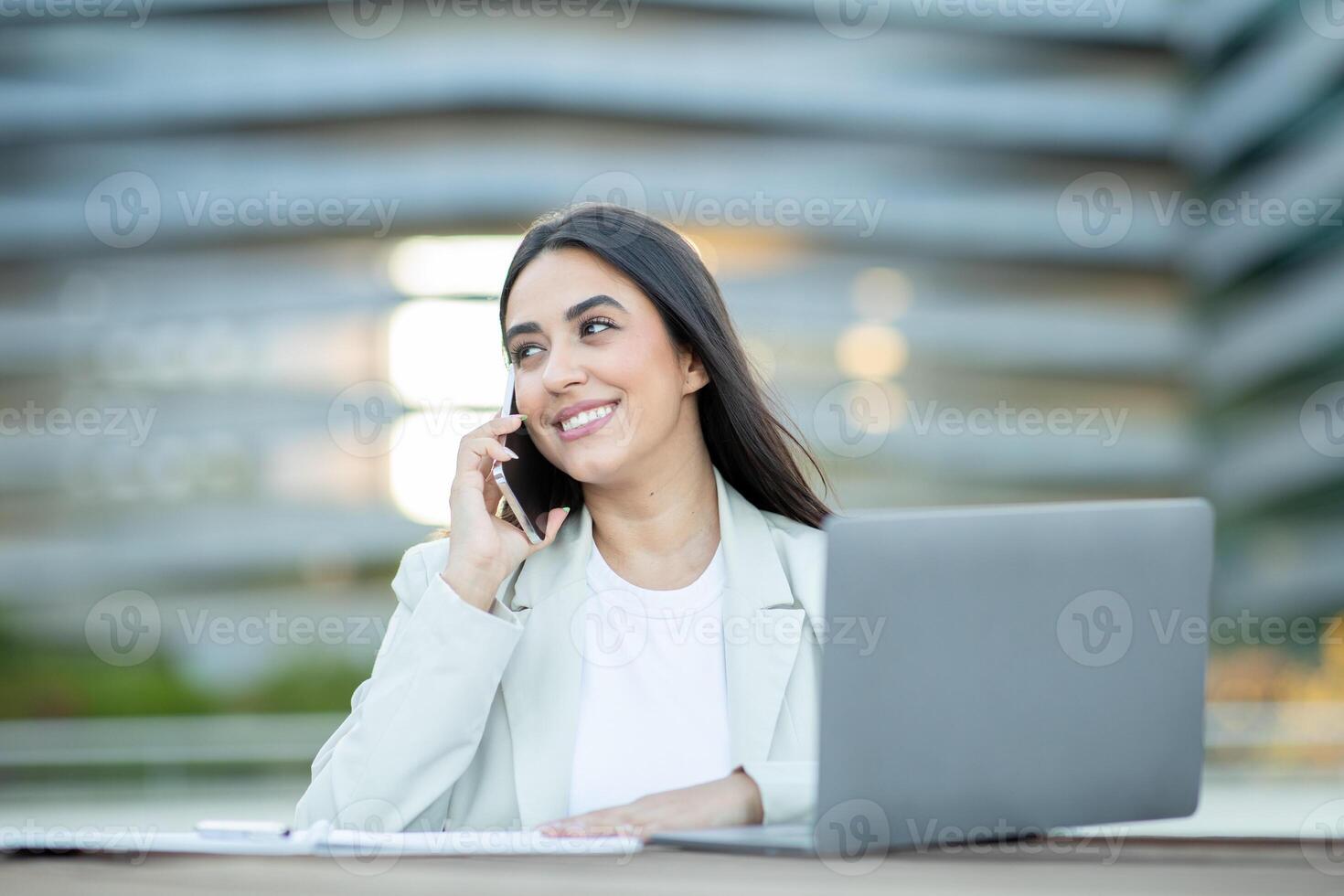 A young woman in a white blazer smiles while talking on her phone, with a laptop in front of her. She is sitting outside in front of a modern building. photo