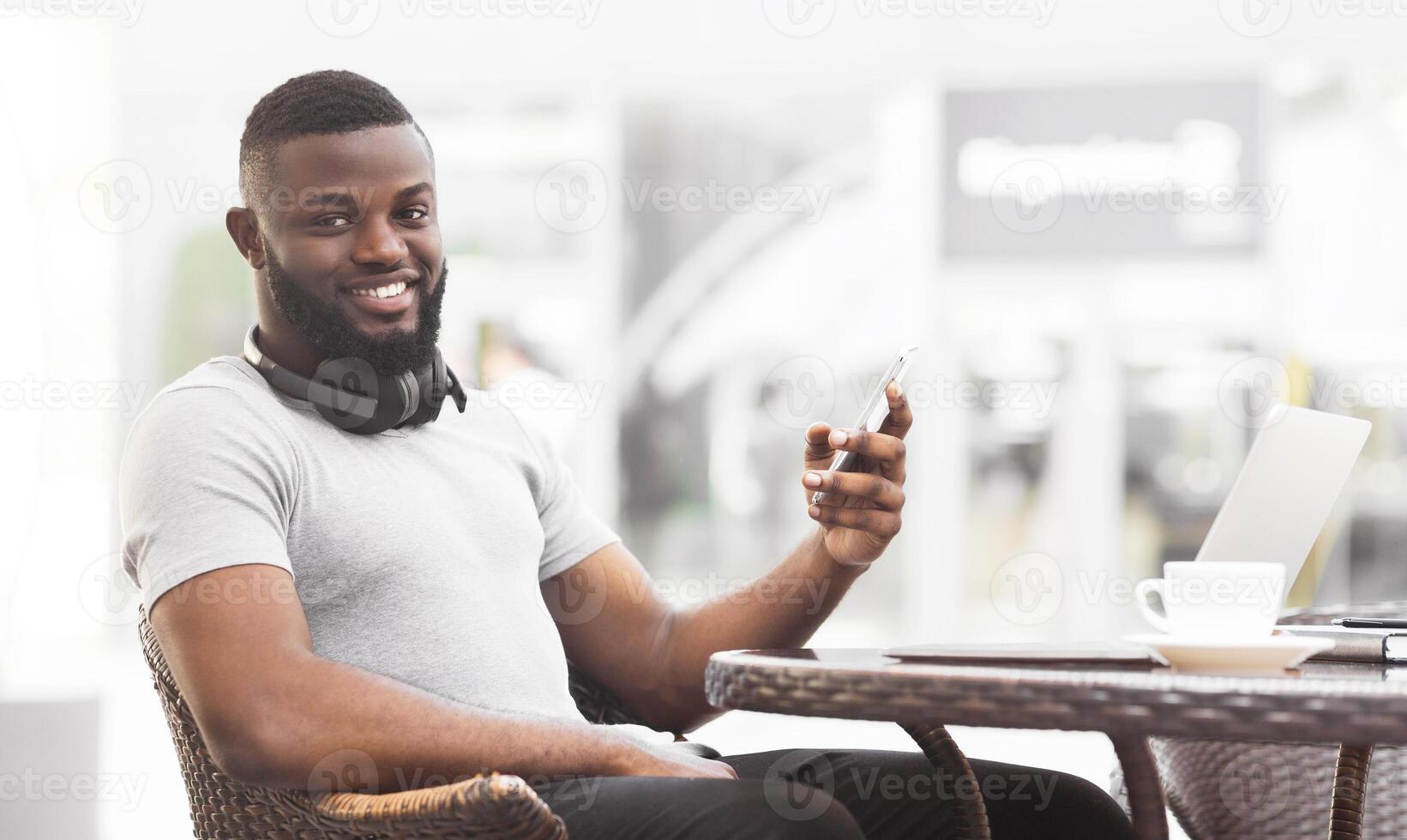 A young man sits at a cafe table with a laptop and a cup of coffee, smiling while looking at his smartphone. photo