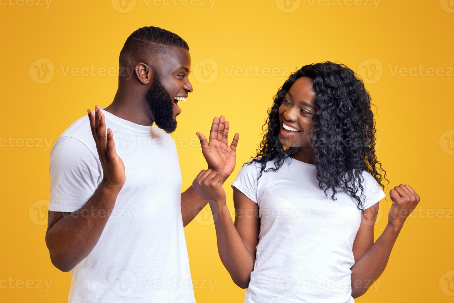 Happy couple in white t-shirts celebrating in front of yellow background with wide smiles and raised hands, looking excited about their achievement photo