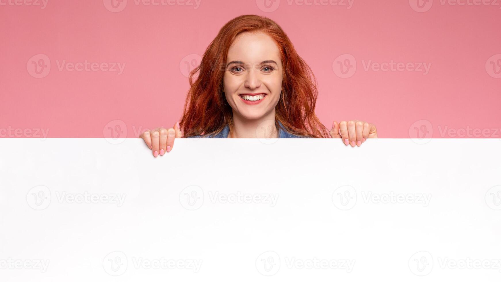 A red-haired woman is holding a white board in her hands. She is looking at the board with a focused expression, possibly in a teaching or presentation setting photo
