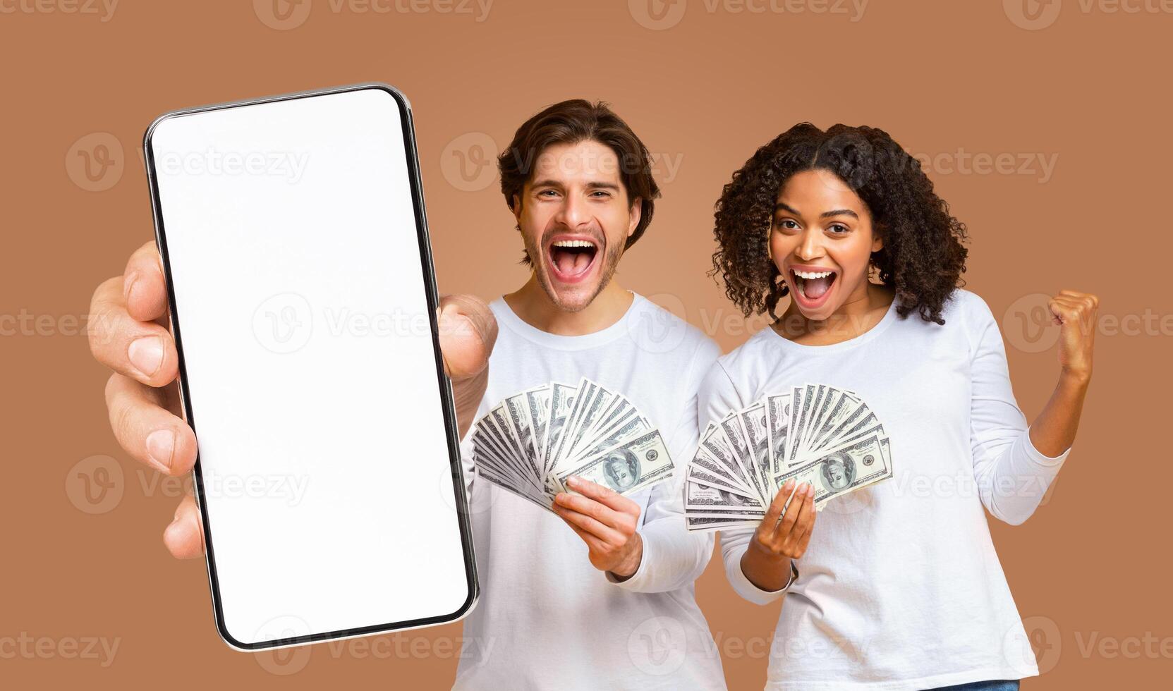 A young man and woman are shown smiling and excited while holding cash in their hands. The man is pointing towards a smartphone with a blank white screen being held in front of him. photo