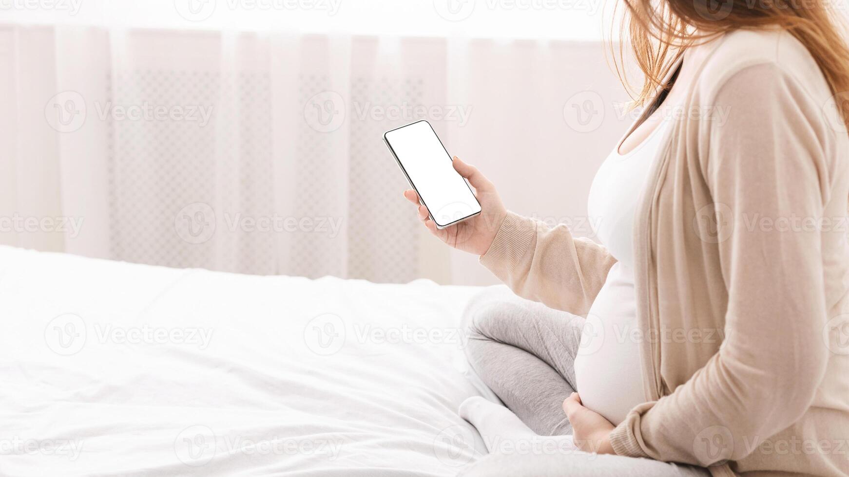 Pregnant woman sitting on bed, using smartphone to find information before birth and child rearing after birth, empty space photo