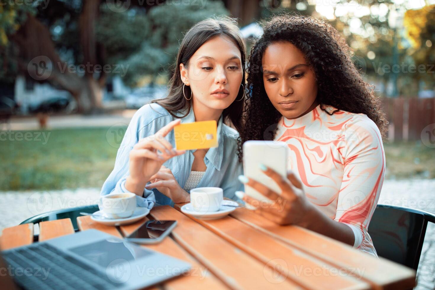 Two young women, one with long dark hair and the other with curly dark hair, sit at a table outside a cafe, checking their phone and credit card. There are two cups of coffee and laptop on the table. photo