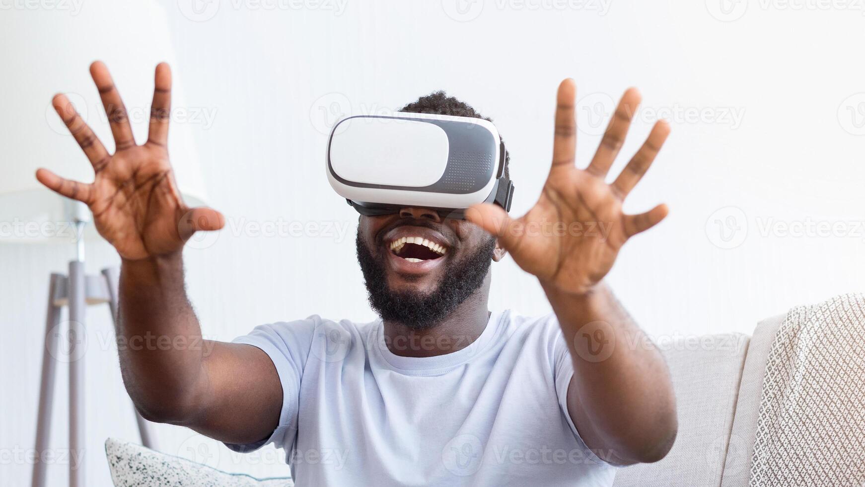 African American man wearing a VR headset is seated on a couch, both hands outstretched, as he laughs in delight at something in the virtual world. photo