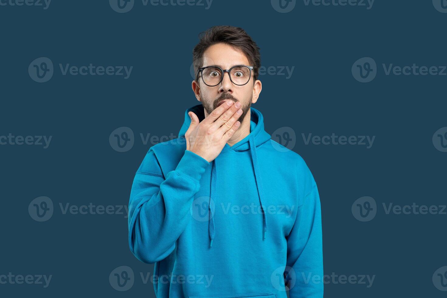 A young man stands against a dark background, wearing a blue hoodie and glasses. He looks surprised, with his hand covering his mouth, expressing shock or disbelief at something unexpected. photo