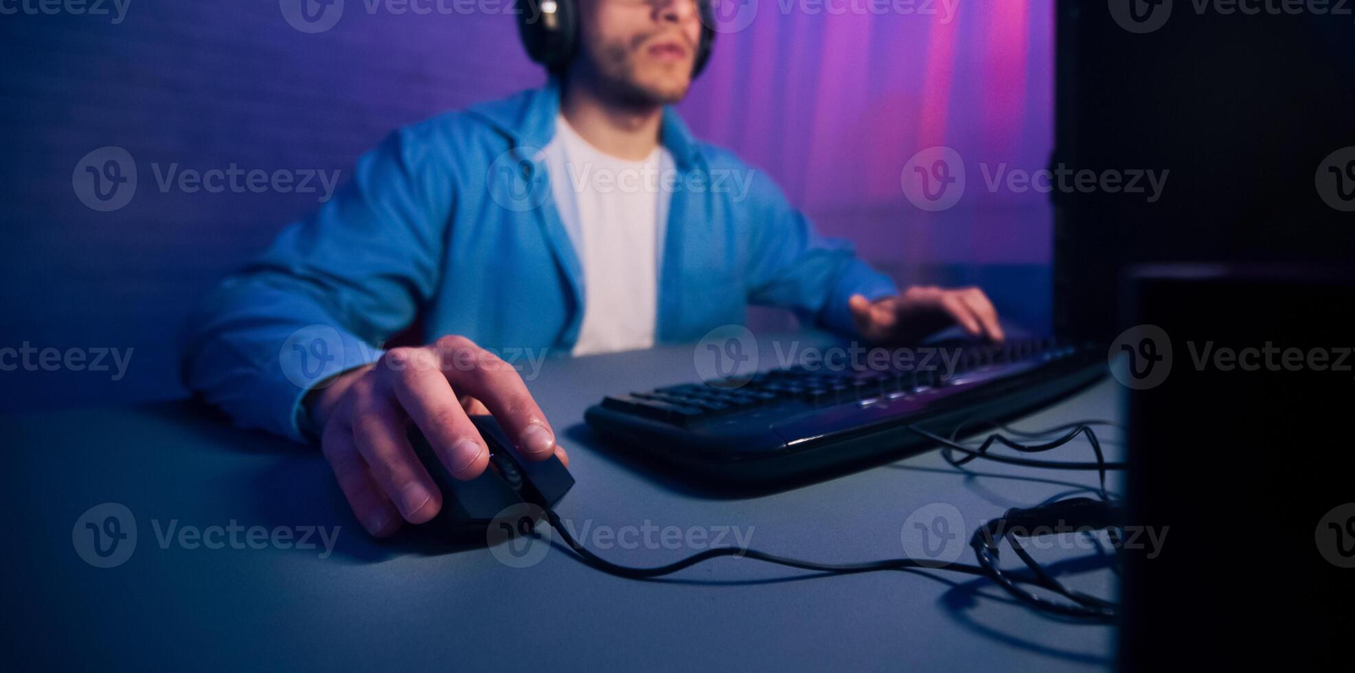 A mans hand rests on a computer mouse, with the keyboard in the background. The man is wearing a blue hoodie and headphones, suggesting he is gaming, cropped photo