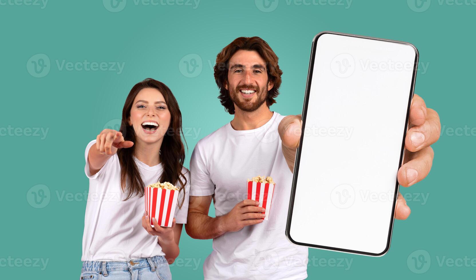 A smiling couple enjoying a movie on a smartphone. They are both holding red and white striped containers of popcorn, and the man is holding the phone forward for the viewer to see photo
