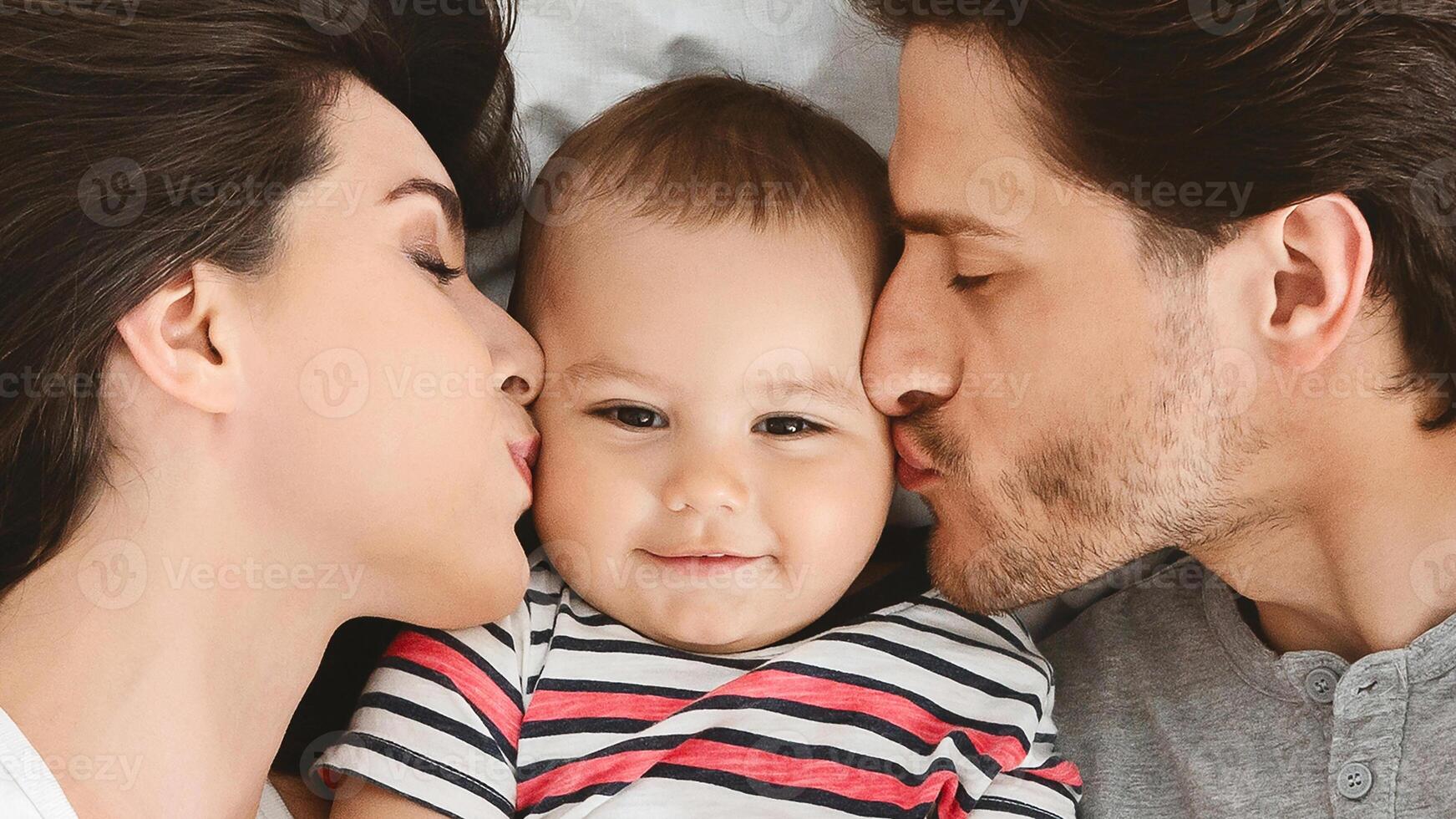 A happy baby lies in the middle of his parents, who are both kissing him on the cheek. The baby is smiling, and the parents both have their eyes closed, closeup photo