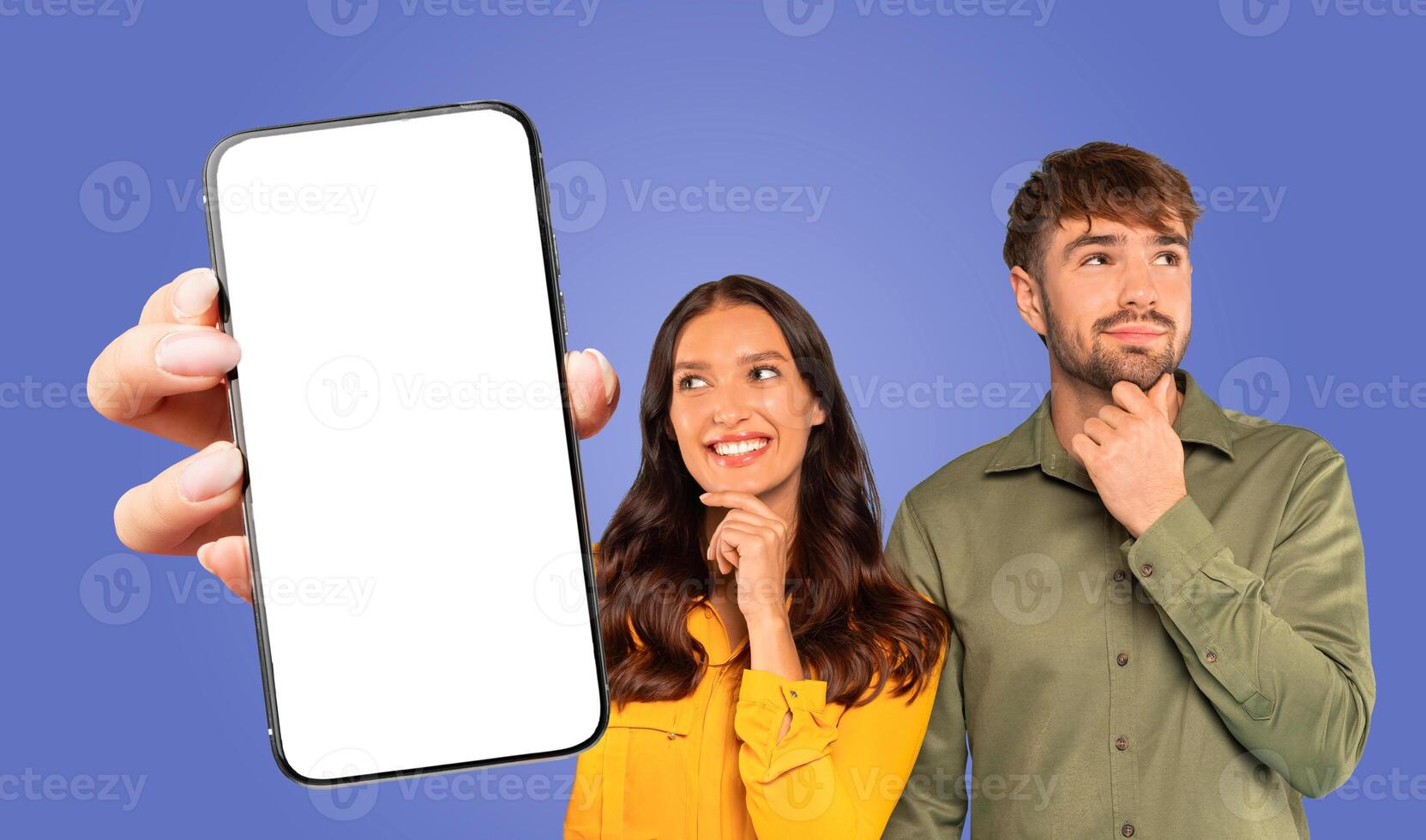 A woman holds a smartphone with a blank screen in front of a blue backdrop. She is smiling and looking at the screen. The man beside her is looking upwards while holding his chin photo
