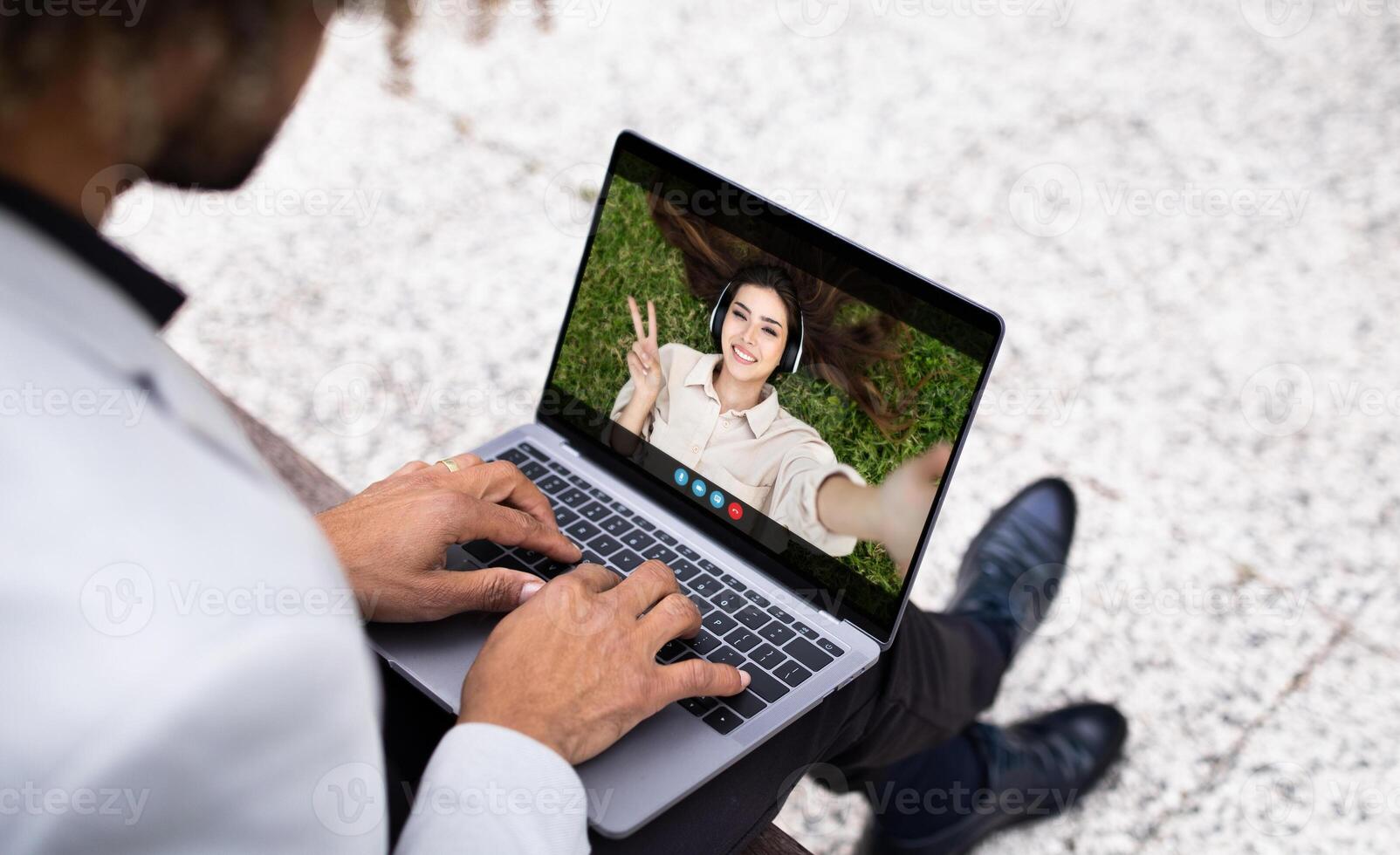 Black man sits outside on a bench, using a laptop to chat with a woman. The woman is smiling and giving a peace sign. The man is typing on the laptop keyboard. photo