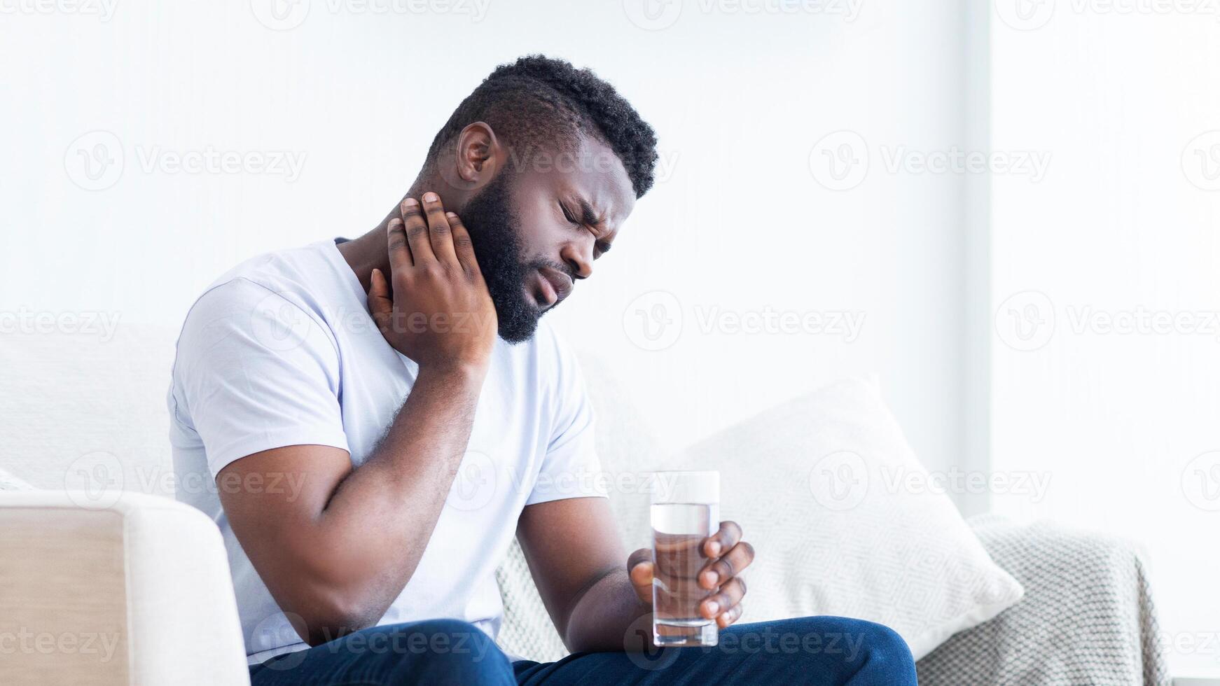Black man with a beard sits on a couch, holding a glass of water in his right hand and touching his neck with his left hand, appearing to have a sore throat, copy space photo