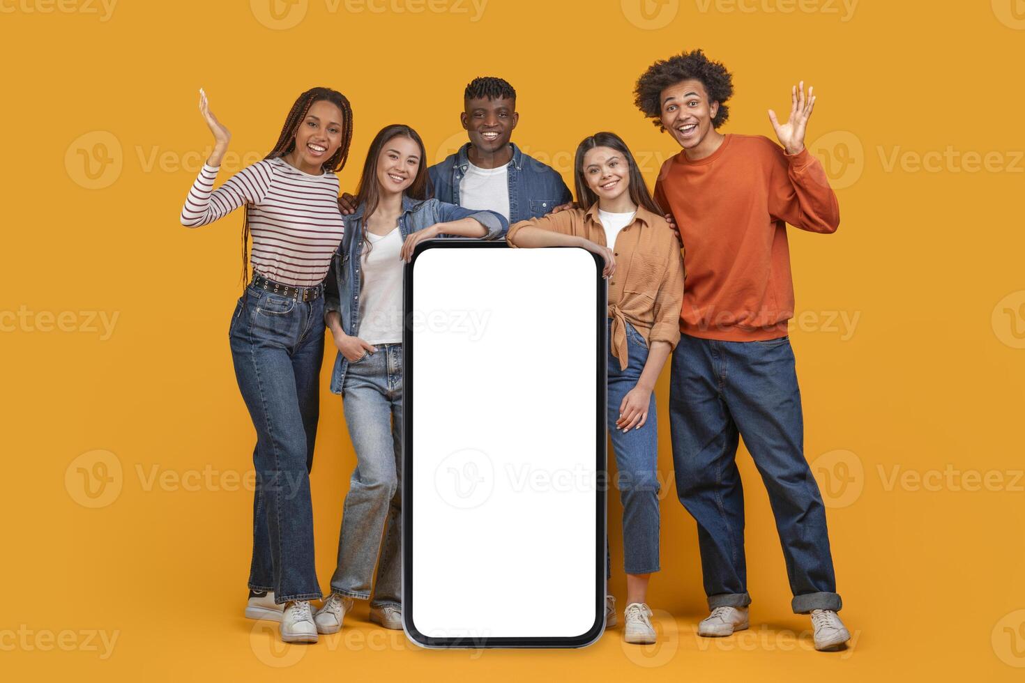 Five happy young friends stand in front of a vibrant orange background, holding a large smartphone with a blank white screen. They are smiling and laughing photo