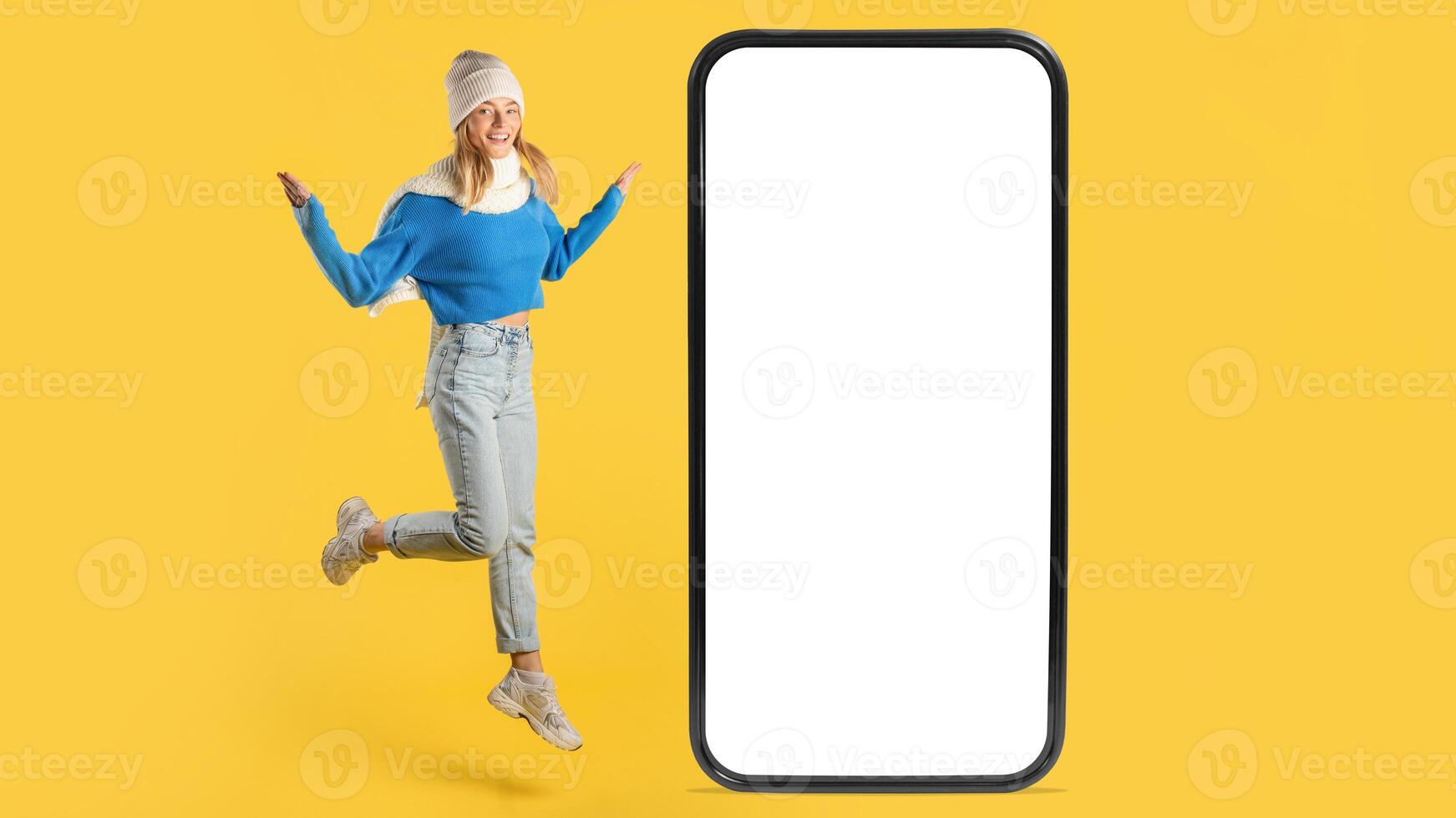 A cheerful young woman in a blue sweater and jeans joyfully jumps next to an oversized smartphone with a blank screen. The vibrant yellow background adds a lively touch to the scene. photo
