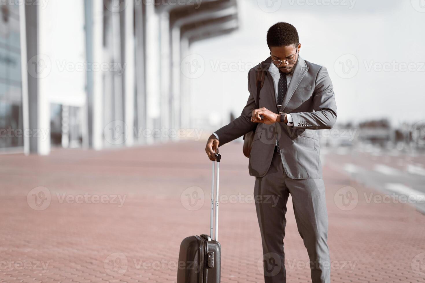 Black businessman checking time on his watch with rolling suitcase outside a modern building, preparing for a business trip. photo