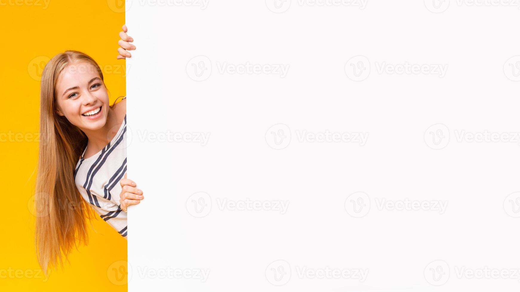 Excited smiling girl looking from behind blank advertising board over yellow background, panorama photo