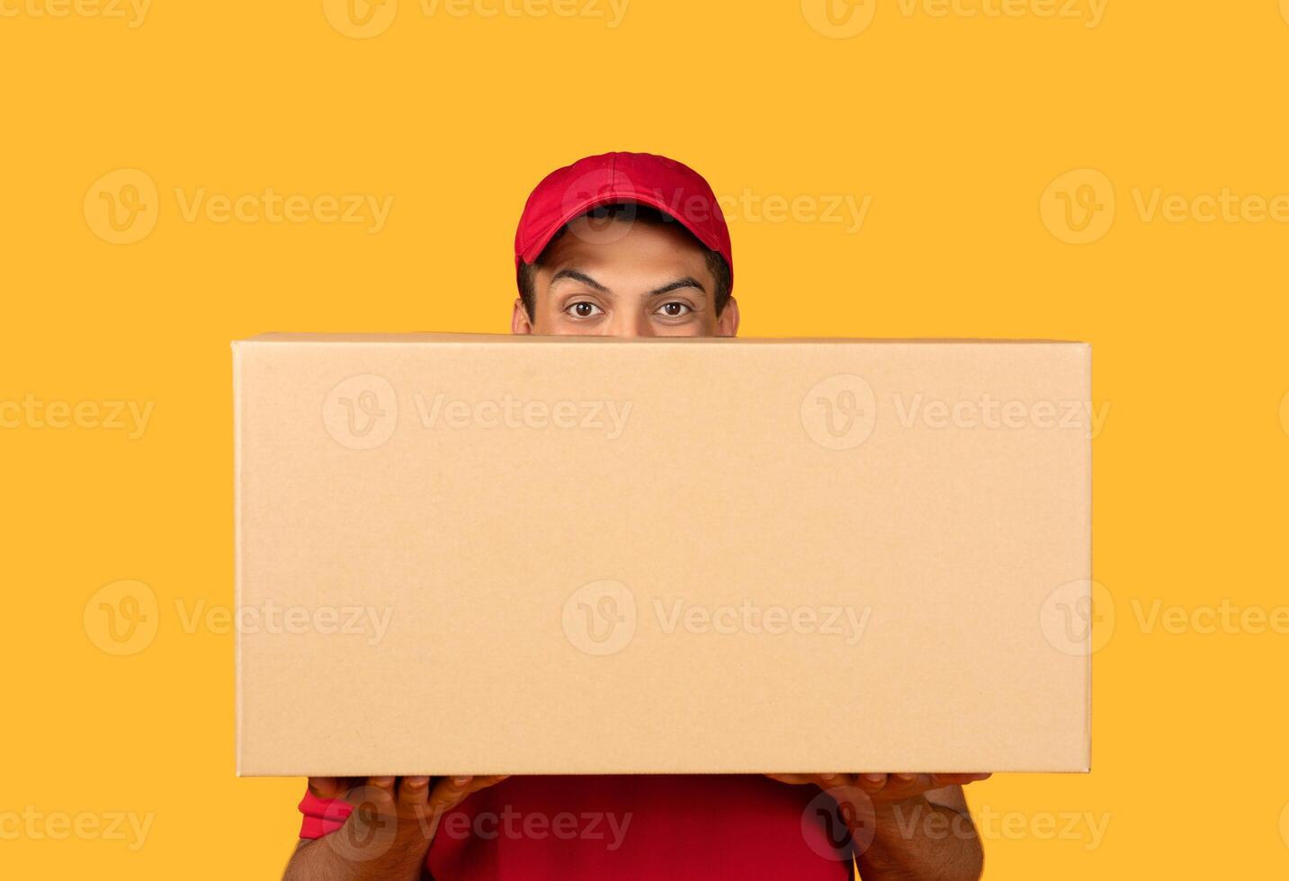 A delivery man wearing a red uniform and cap is holding a large cardboard box. The background is a solid, bright yellow, making the scene vibrant and eye-catching. photo