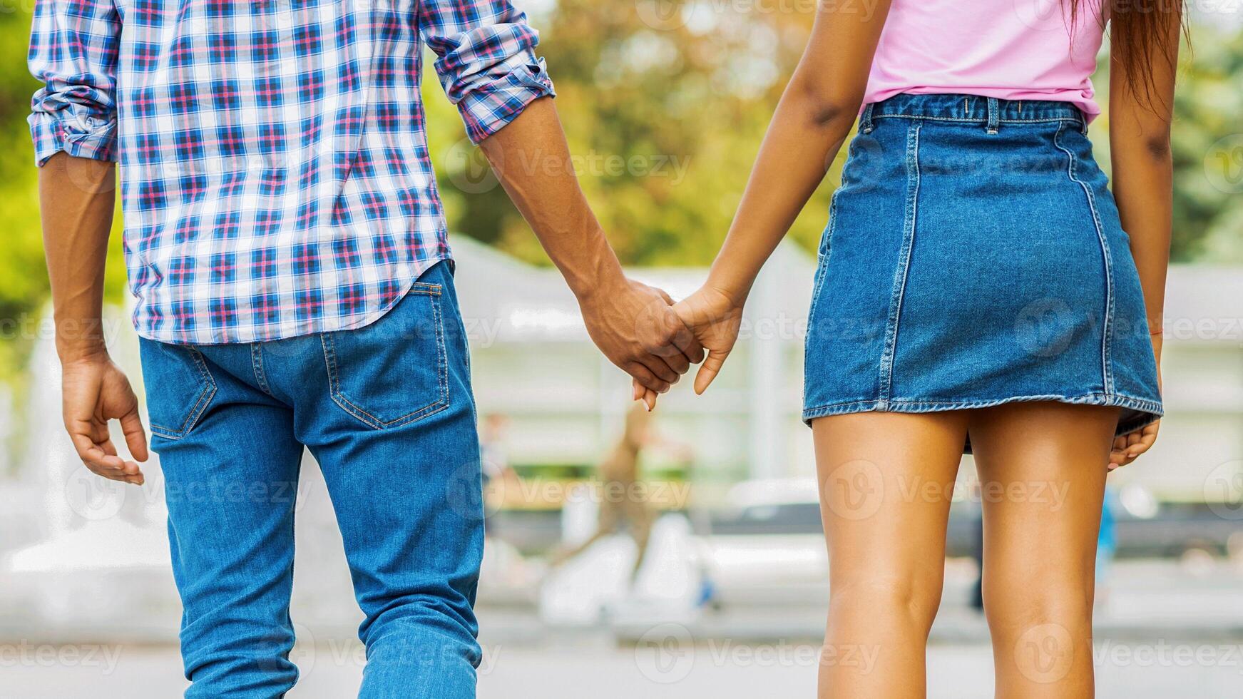 A couple walks hand in hand, enjoying a day out in the summer. Guy is wearing a blue and white plaid shirt and blue jeans, while the girl is wearing a pink shirt and a denim skirt, back view, cropped photo