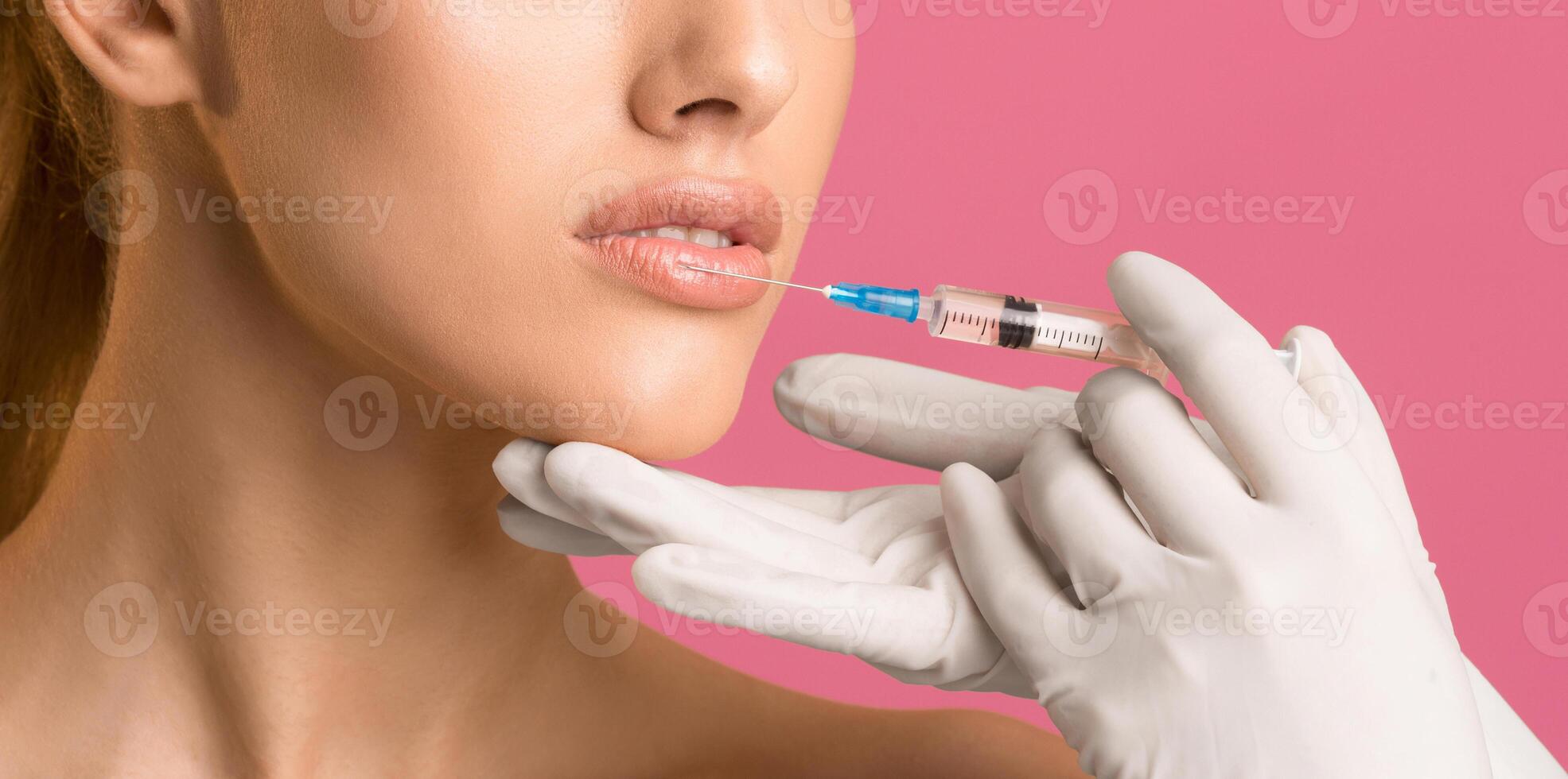 A close-up image of a woman receiving a lip injection. Her lips are parted slightly, and a gloved hand is holding a syringe near her mouth. The background is a vibrant pink. photo