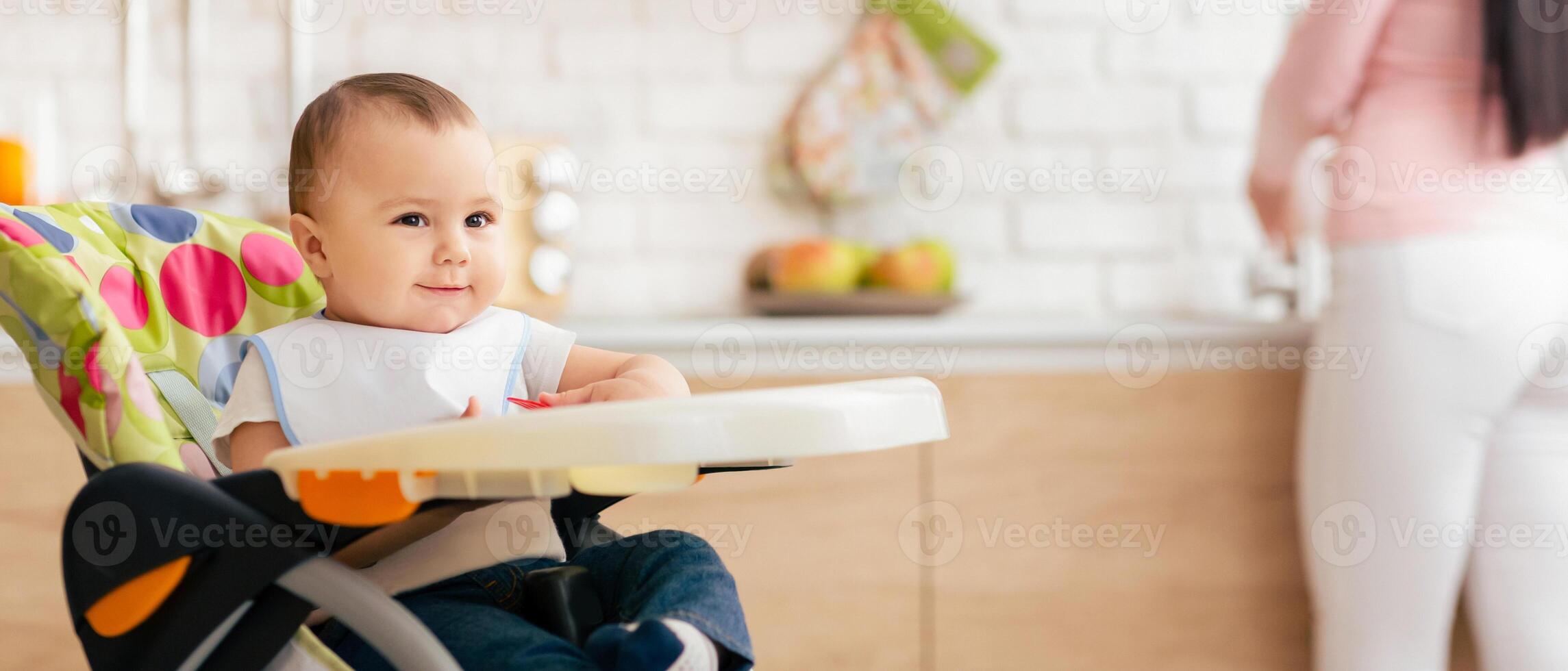 A baby is comfortably sitting in a colorful high chair in a bright, modern kitchen. In the blurred background, the mother is seen preparing food on a countertop, indicating a typical morning routine. photo