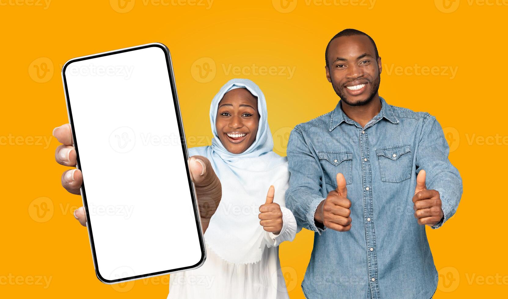 Black woman wearing a headscarf and a man, are holding a smartphone with a blank screen and giving thumbs up. They are smiling and looking directly at the camera photo