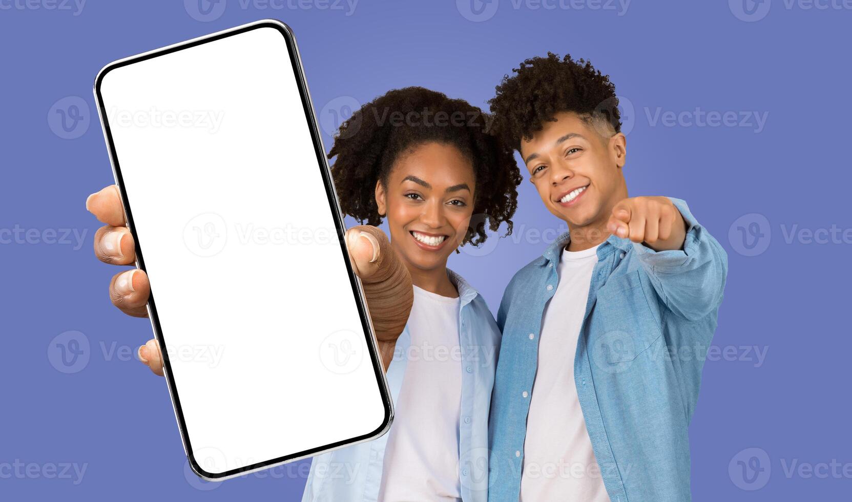 A young black woman and man are standing side-by-side, smiling and pointing at the camera. They are holding a smartphone with a blank screen in front of them. The background is a light blue color. photo