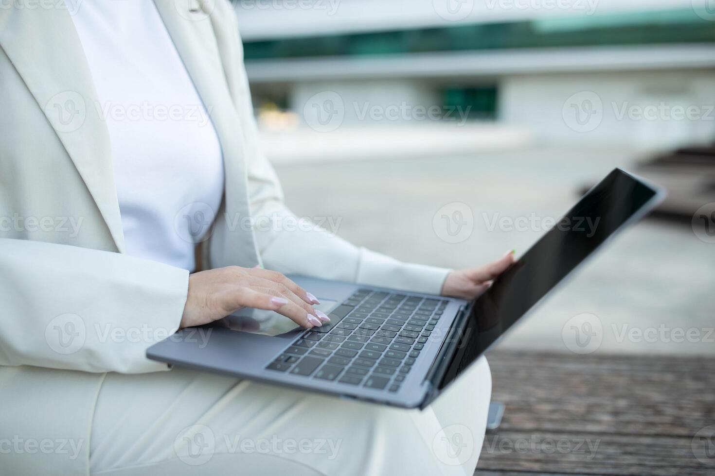 A woman wearing a white suit is sitting on a bench outside. She is using a laptop and appears to be working or browsing the internet. The background is blurred, suggesting a modern urban environment. photo