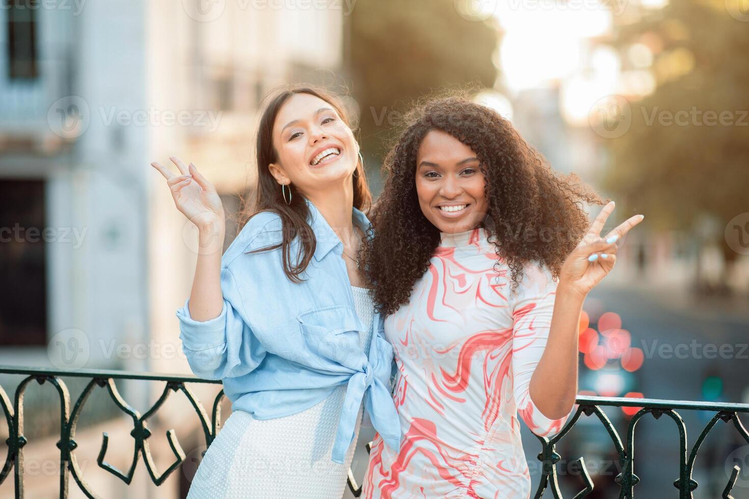Two women are standing on a city street, smiling and posing with peace signs. They appear to be enjoying their time together as they stand in front of a wrought iron fence. photo