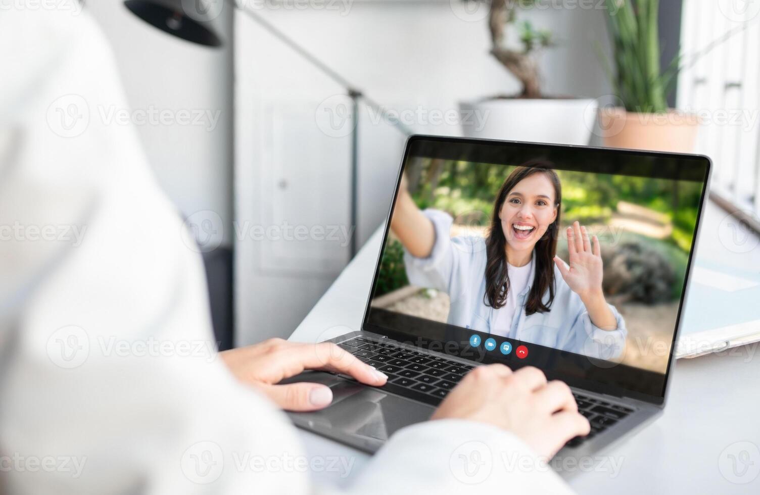 A woman is sitting at a desk in an office setting and chatting with a person on her laptop. The person on the other end of the call is waving to the camera. photo