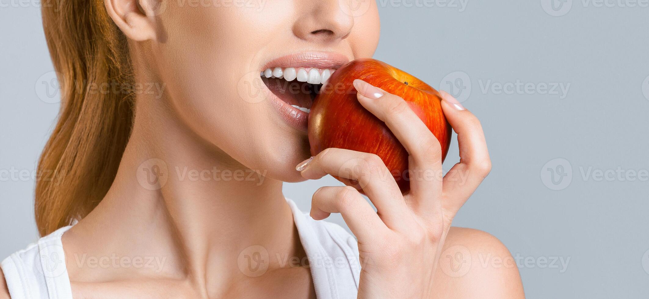 A close-up shot of a woman taking a bite out of a red apple. She is wearing a white tank top and her hair is pulled back in a ponytail. The photo is taken against a light grey background.