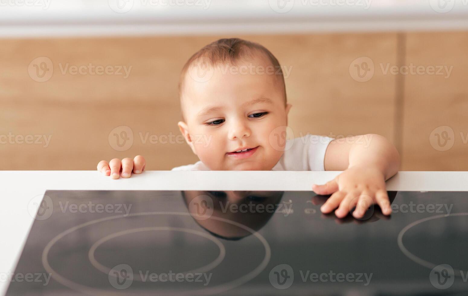 A curious baby in a white shirt reaches towards an induction stove in a modern, well-lit kitchen. The baby appears fascinated and intrigued by the shiny surface. photo
