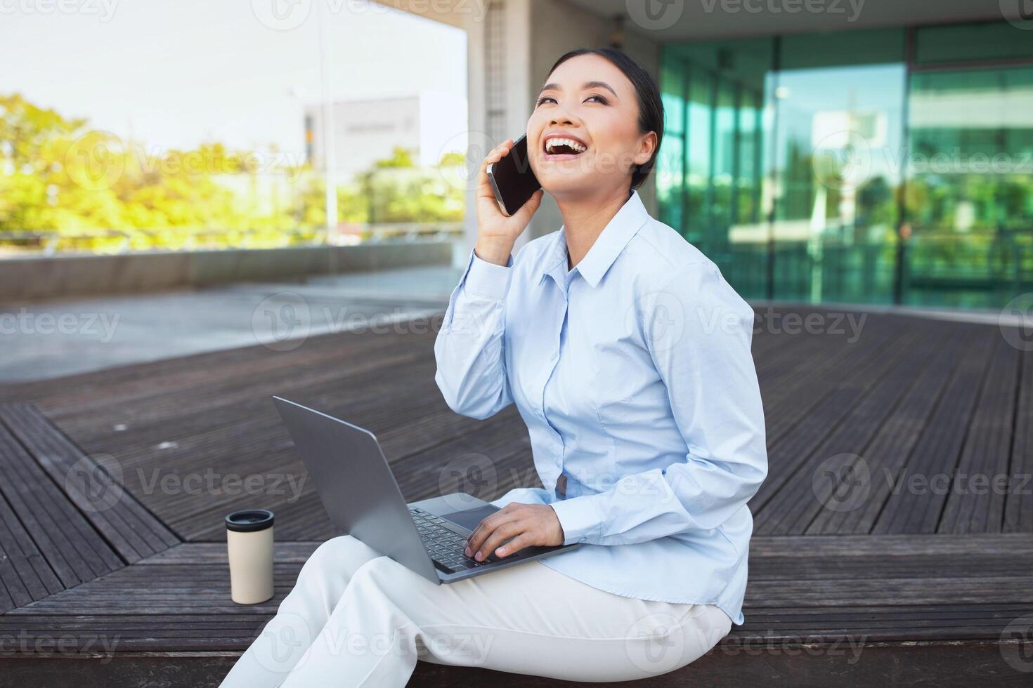 Asian woman is seated on a bench, engaged in conversation on her phone, holding computer on her lap. She appears focused, possibly discussing important matters photo