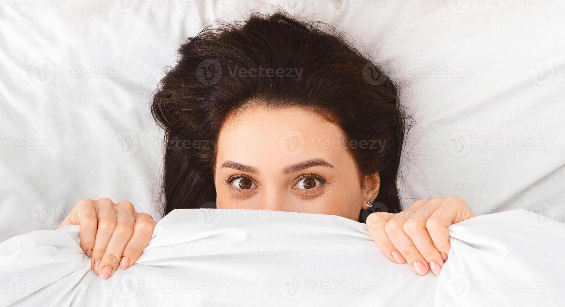 Closeup of woman is lying in bed, covering herself with a blanket, partially hiding her face. She appears to be seeking comfort or privacy in a cozy setting. photo