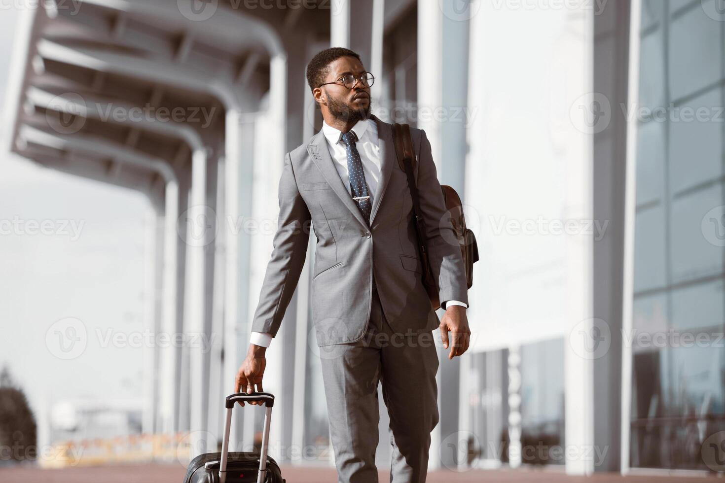 African American businessman walking with rolling suitcase outside a modern building, ready for a business trip or travel. photo