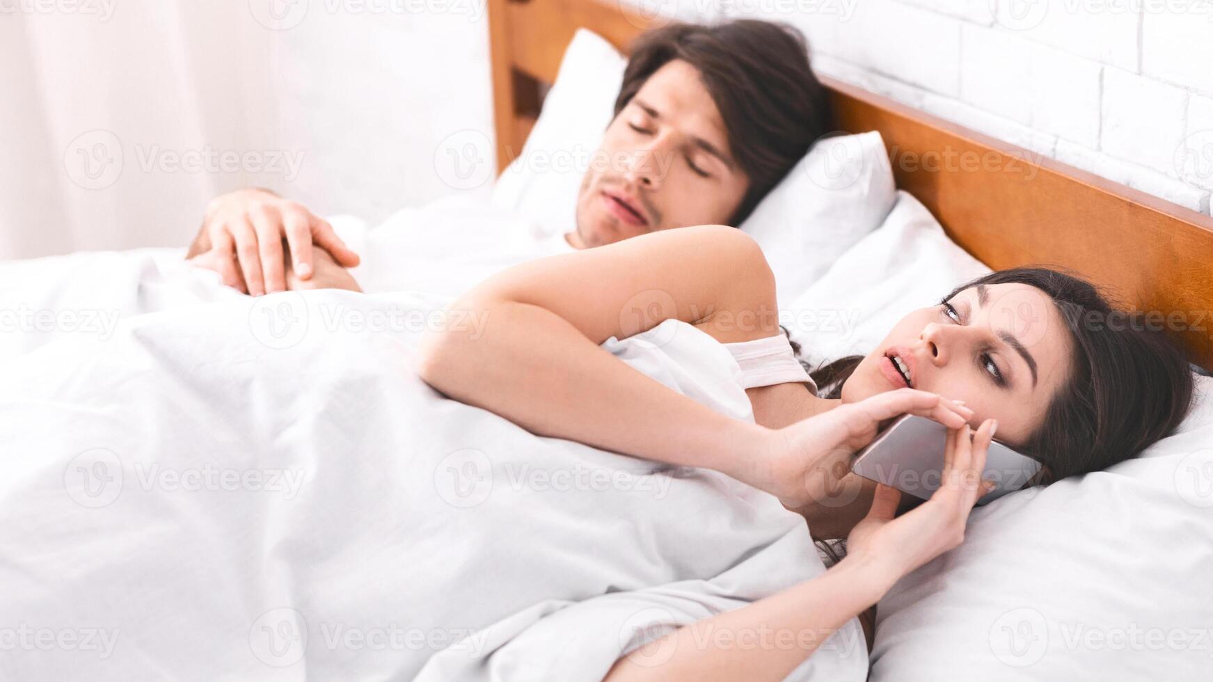 A woman lies in bed, talking on her phone while her partner sleeps soundly beside her. The woman expression suggests she is engaged in a private conversation. photo