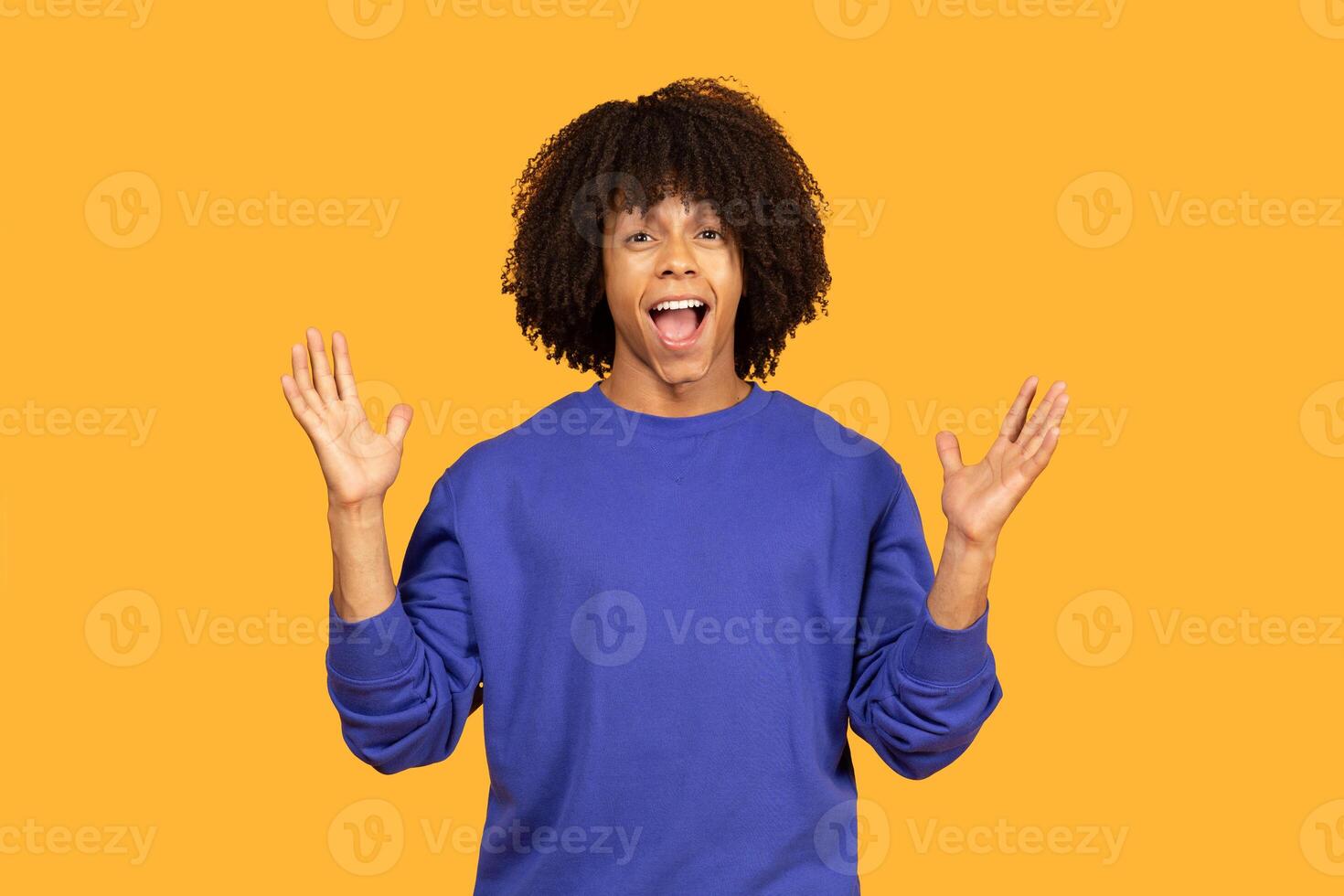 Young man with curly hair wearing a blue sweatshirt and standing in front of a yellow background. He has a surprised expression on his face and is holding both of his hands up in the air. photo