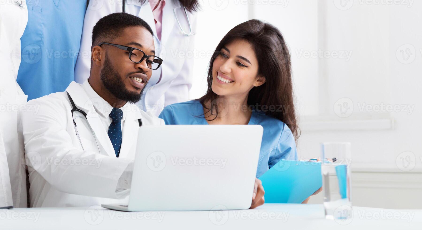 Team of interns and practitioners examining medical reports at hospital, using laptop photo