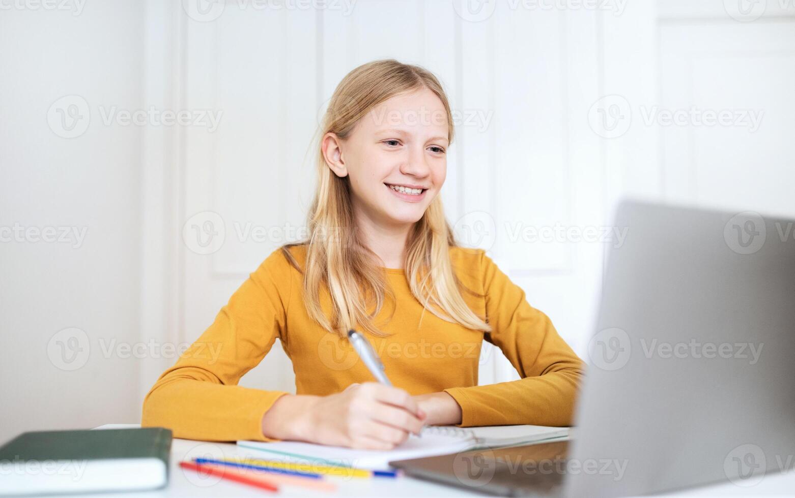 A teen girl with long blonde hair sits at a desk in front of a laptop computer. She is writing in a notebook with a pen and smiling. There are colorful pencils and a closed book on the desk. photo