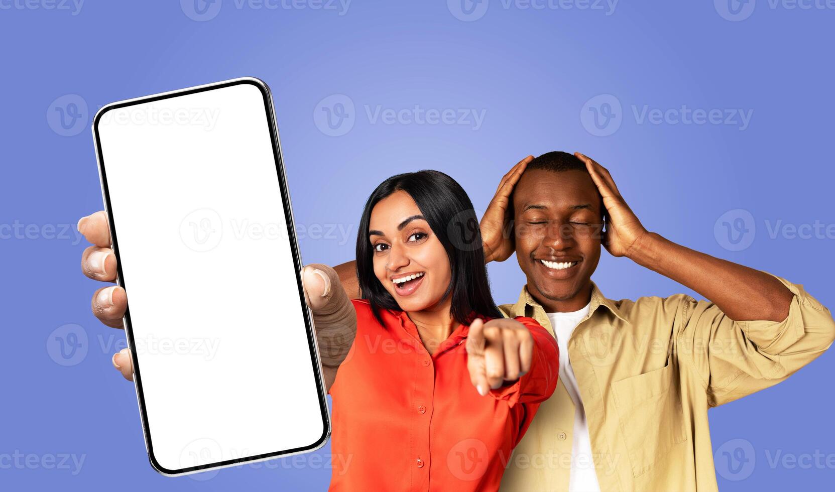 Indian woman with long dark hair and a red shirt holds a smartphone with a blank white screen in her left hand and points at it with her right hand. Behind her, a black man holding his hands to head. photo