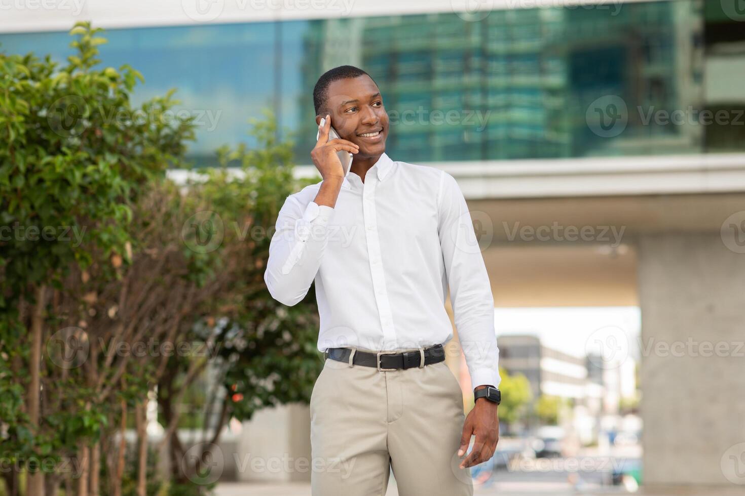 African American man in a white dress shirt and khaki pants stands on a city street while talking on his mobile phone. He has a smile on his face and is looking off to the side. photo