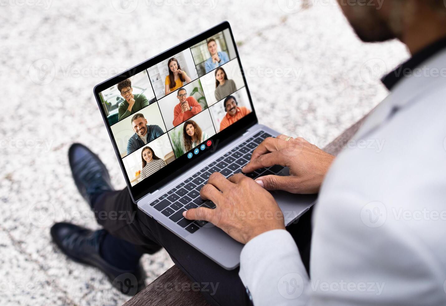 Black man sits on a bench outdoors, using his laptop to participate in a conference meeting. The screen displays a grid of nine participants in various locations, all connected remotely photo