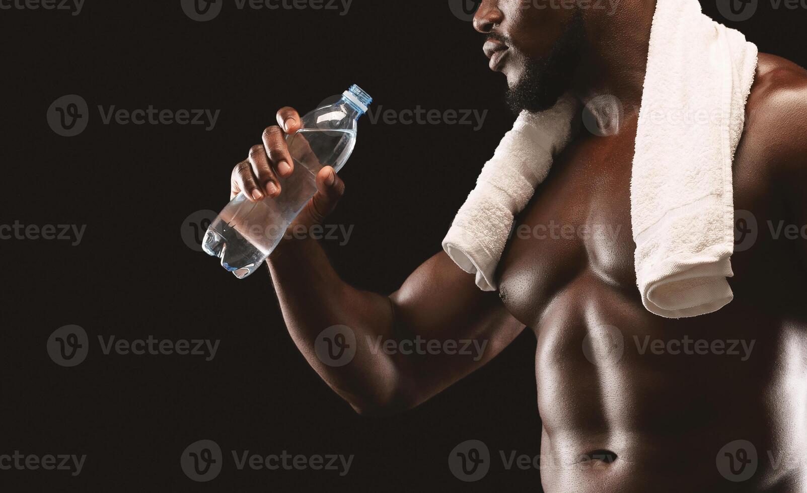 A muscular man, with a towel draped over his shoulders, drinks water from a plastic bottle after a workout. photo